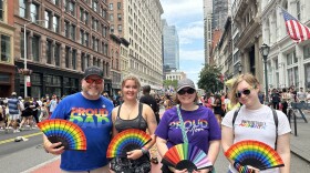 Dan, Ava, Sonya, and Zo Moyle stand next to each other on a street in New York City, during the 2023 Pride March. They're all carrying pride themed rainbow hand held fans, with other pride related swag also being worn by the group like Dan Moyle's "Proud Dad" pride colored t-shirt. A crowd of people walk and stand around them as the tall buildings of New York sit on both sides of them. 