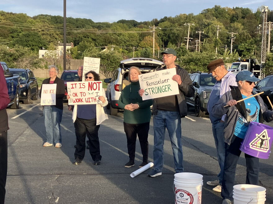 Several protesters are standing in a row in a parking lot. Most of them are holding posters, some of which say "It is NOT ok to with ICE" and "Immigrants make Rensselaer STRONGER." One woman is holding drum sticks and has a plastic bucket hanging from a strap around her neck. 