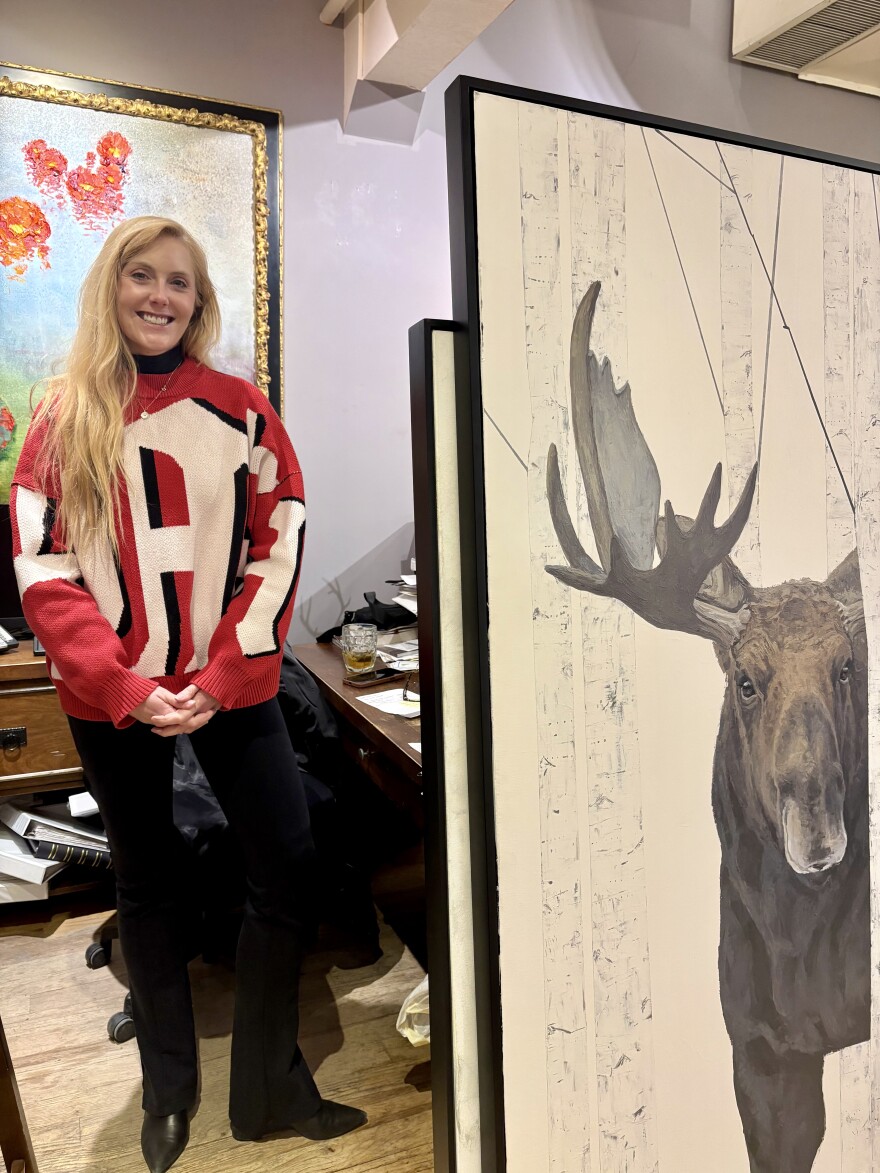 A woman in a red sweater poses in next to a painting of a moose.
