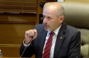A man in a suit and red striped tie sits at a conference table, speaking into a microphone. He appears to be in a formal meeting room with a wooden door and leather chair visible in the background.