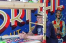 A smiling man leans onto a painting scaffold positioned in front of a colorful mural.