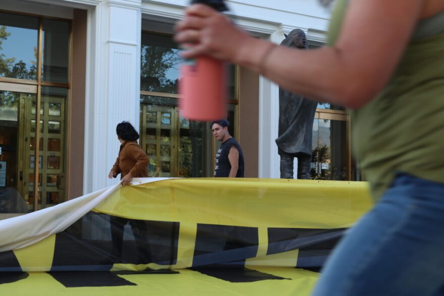 Members of WildEarth Guardians and New Mexico Youth United Against Climate Crisis Action (YUCCA) unfurl a banner on the grounds before the Roundhouse on the evening of Oct. 7, 2025, protesting Exxon-Mobil’s 2025 sponsorship of the Albuquerque International Balloon Fiesta. (Danielle Prokop / Source NM)