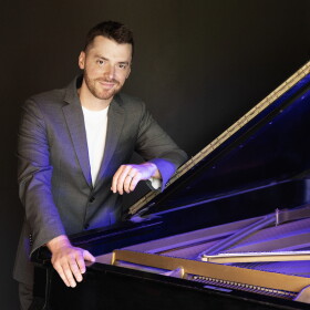 Peter Dugan stands leaning against an open piano