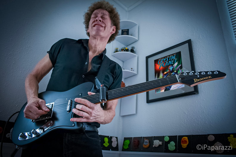 Musician Ned Evett stands in a living room playing a guitar.