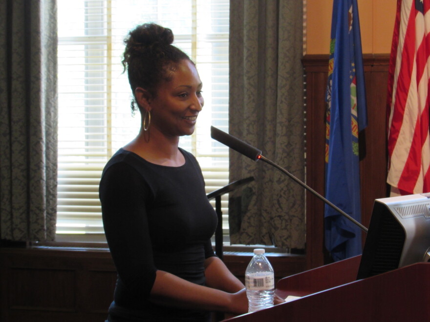 A young woman in a black top stands at a lectern with a microphone. She is smiling.