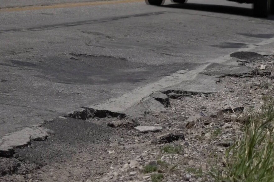 A badly maintained side of a road is pictured. There are many pot holes and crumbling asphalt. 