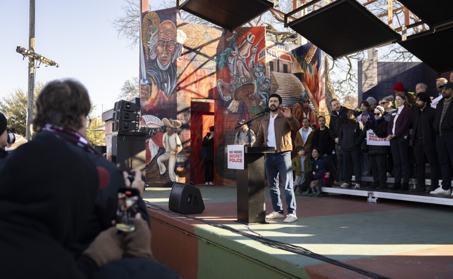 A man stands on stage at a podium with others in the background and speaks to a crowd. 