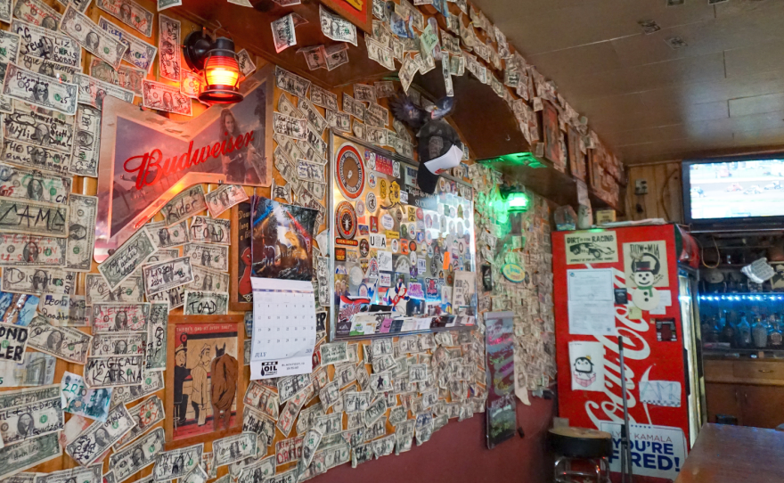 A store wall covered stickers, signs, and dollar bills that people have written on.