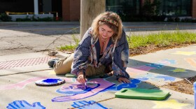 Delia Kerr-Dennhardt painting two colorful trapezists on the sidewalk near the Bloomington Public Library.