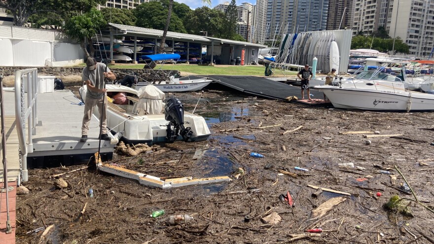A Waikiki Yacht Club employee (left) adjusts a makeshift wooden tool meant to corral debris at the Ala Wai Boat Harbor. Volunteers Jacob Stone and Jeovanney Gutierrez (right) wait by a boat they will use to drag the tool around and collect debris. (Oct. 29, 2024).