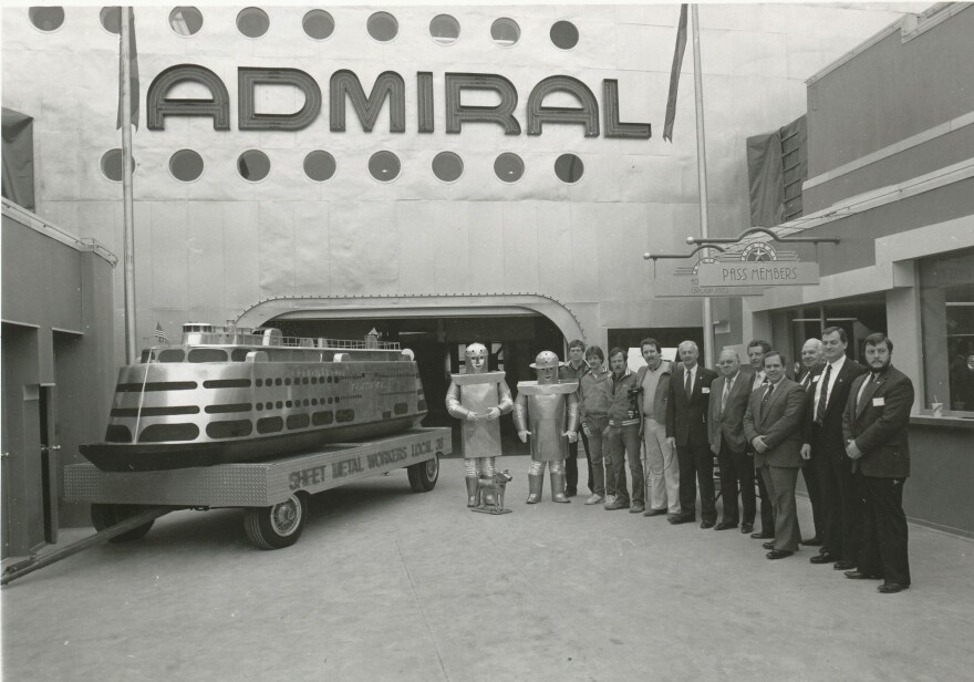 A black and white photo shows the SS Admiral steamship in the background. A model of the boat is to the left on a wheeled cart. A group of white men, some wearing suits and some wearing work clothes, stand to the right under a sign that reads pass members.