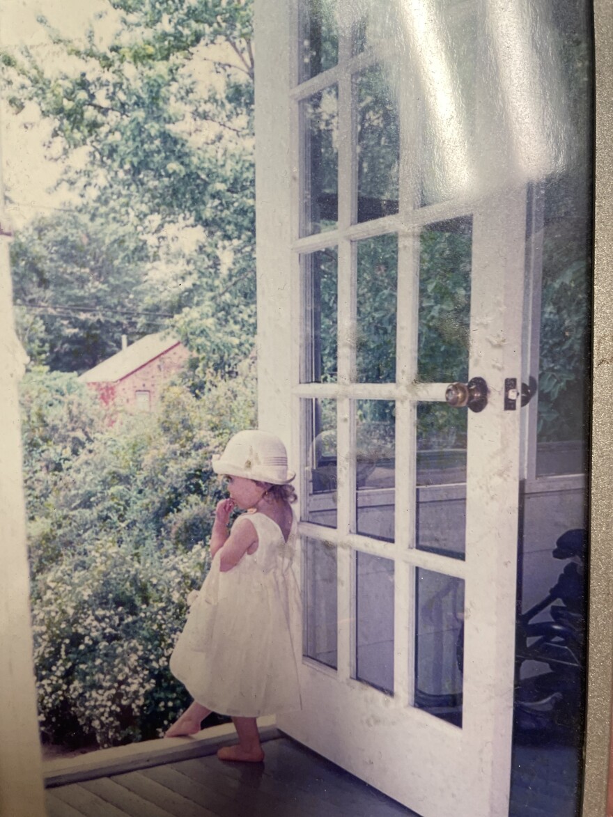 A young girl dressed in white is stepping her foot outside the door of a beautiful garden of white flowers.