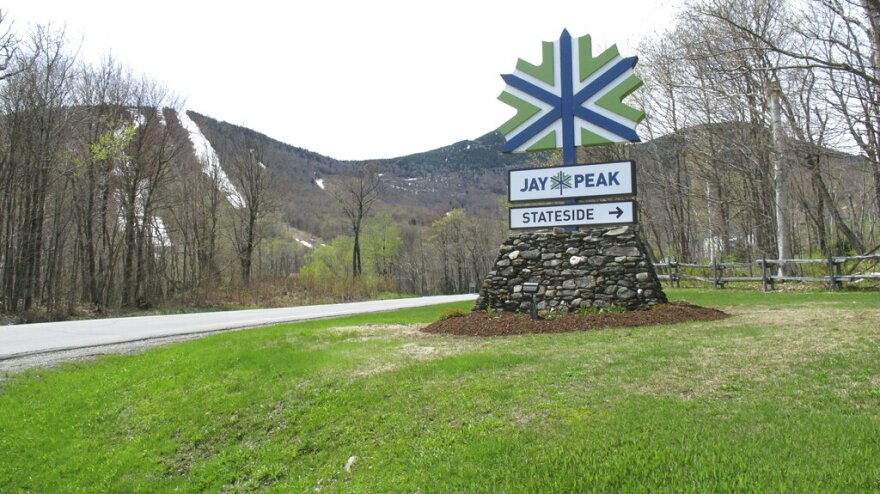 The green and blue leaf-shaped logo of ski resort Jay Peak atop a stone pile foundation sits in a field of green grass.