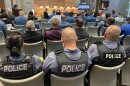 Several uniformed people wearing blue shirts and a black vest sits with their back to the camera in a large meeting room. White letters spelling "POLICE" is printed on the back across the shoulders of their vests.