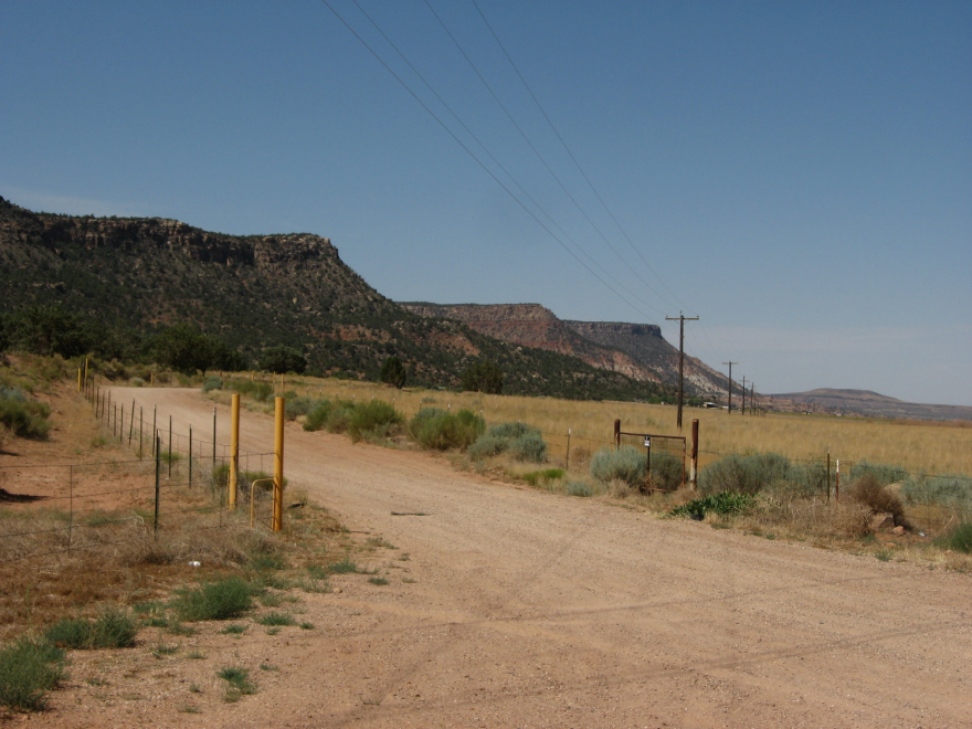 A dirt road near a yellow field with mountains in the near foreground, stretching off into the background.