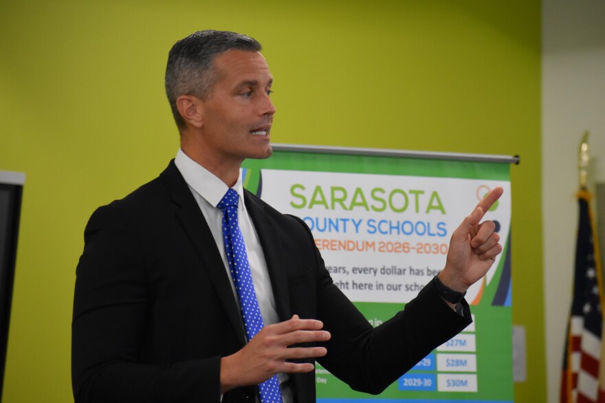 Man with graying hair wearing a dark sut and talking, looking to the right and pointing