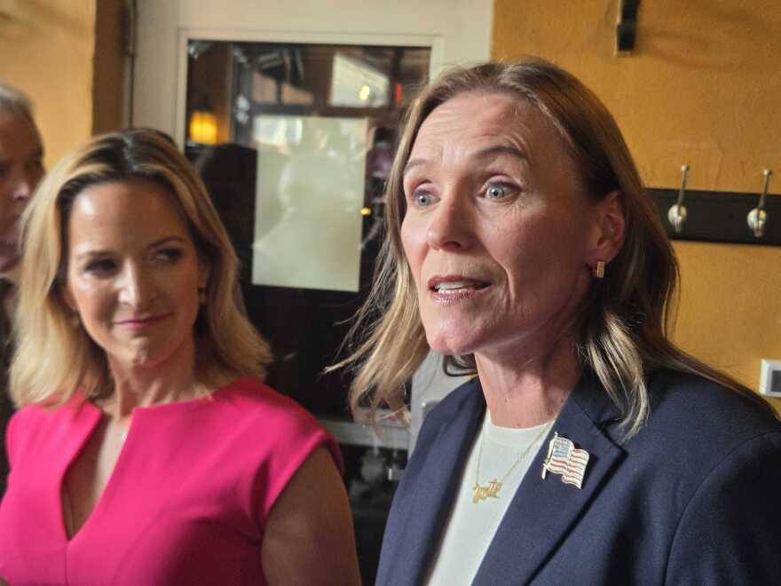 In the foreground, Winnie Brinks wears a navy blazer, American flag pin, and gold necklace. Jocelyn Benson stands beside her in a bright pink dress, smiling and looking toward Brinks. The two women are in a warmly lit room.