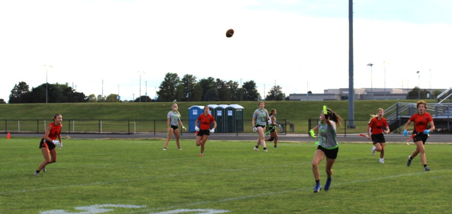High school girls playing flag football on a grass field at a school. Several players are wearing orange and black jerseys. The others are wearing gray and green jerseys. 