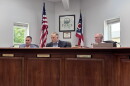 Three men sit behind a large wooden dais, with the Ohio and American flags sitting in the background.