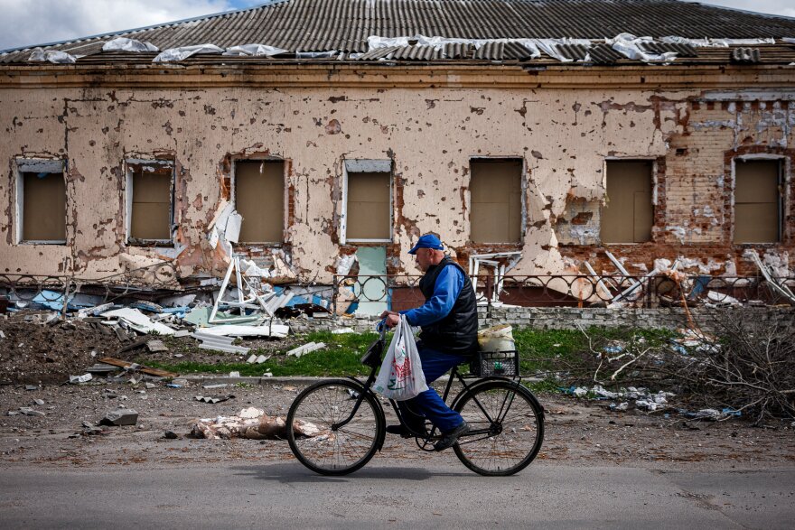A man with a shopping bag rides a bicycle past a heavily damaged house.
