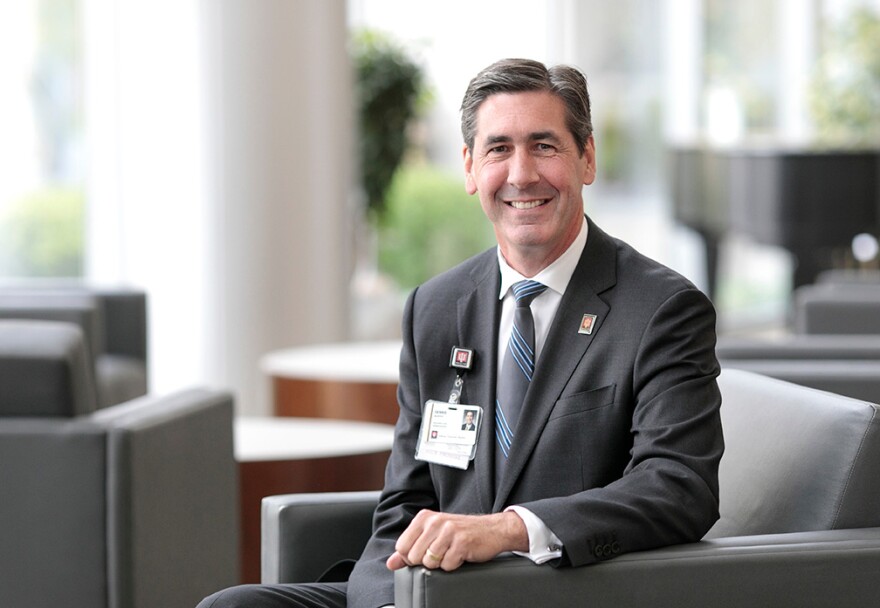 A man smiling at the camera. He is sitting on a black leather chair and wearing a suit and a grey tie with light blue stripes. He haas a small pin on the suit with the letters IU one it.