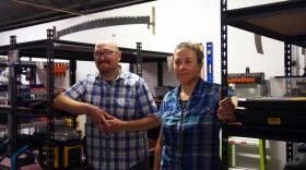 A man and woman in plaid button up shirts stand between two metal shelves, filled with tools. Levels and ladders hang on the white wall behind them, and a long hand saw is hanging at the top of the wall to the top left of the image.