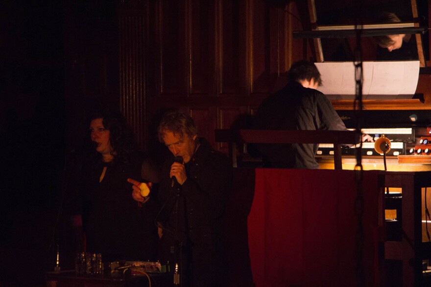 Woman and man singing into microphones, man is holding an apple with a bite out of it, behind them a man plays an organ