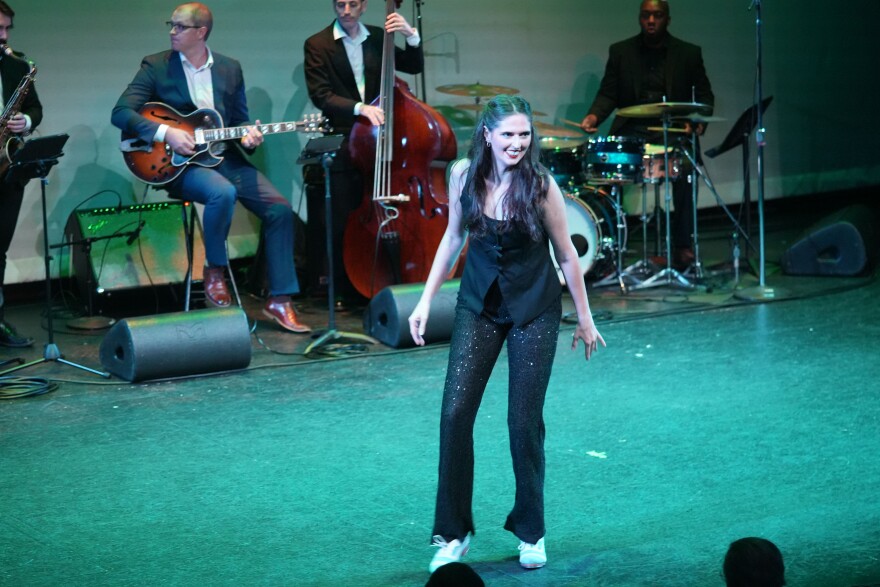 Woman with long brown hair tap dances on stage with band behind her 
