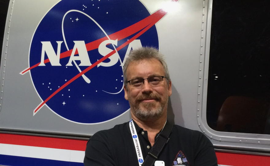 A man with short grey hair and a grey goatee is wearing a black polo shirt and eyeglasses and standing in front of a NASA emblem.