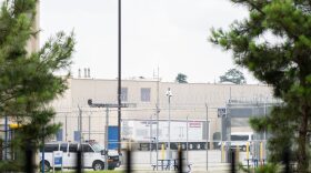 The exterior of a ICE detention center. You can see razor wire atop a chain link fence and a white building.