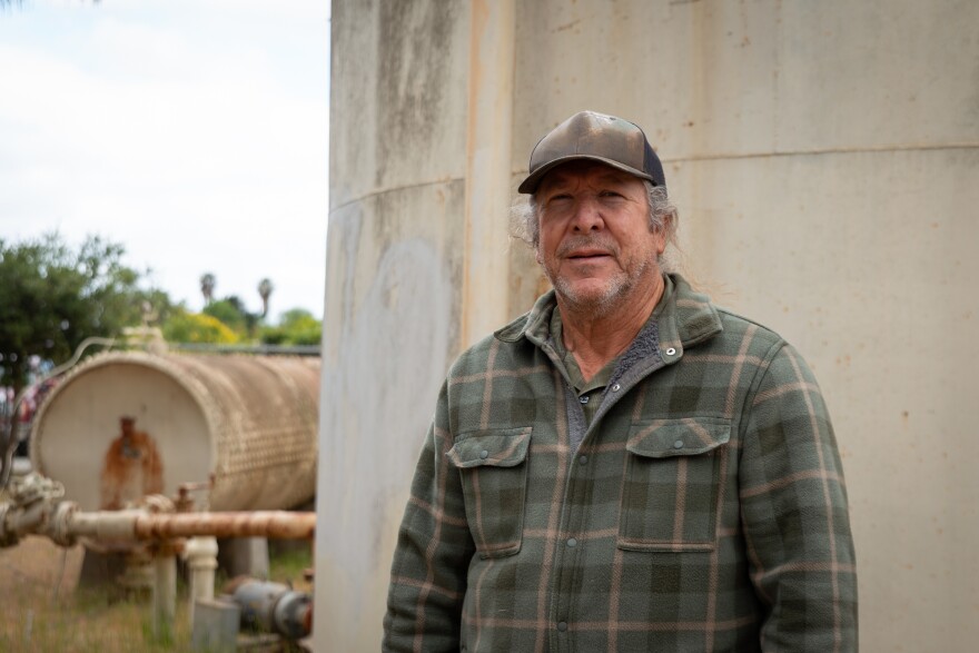 Photo shows a man in a baseball cap and flannel shirt standing in front of an old water system.