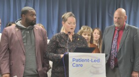 Three people surround a podium with a sign that reads 'patient-led care.' There are other people behind them against a blue curtain.