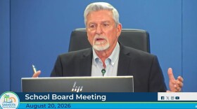 A gray haired man gestures with both hands at the school board dais.