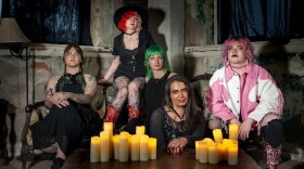 Five people in goth apparel sit on a vintage sofa in front of a low coffee table with many candles on it.