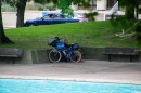A man sits behind a bike with multiple bags strapped to it, avoiding the sun in Freimann Square.