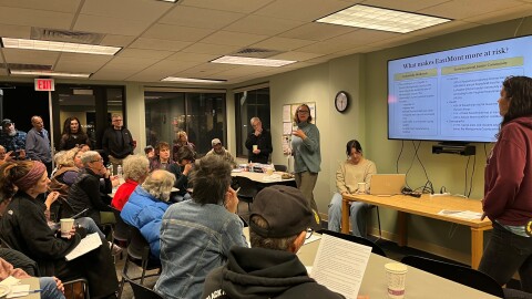 A woman in a blue shirt with chin-length hair and glasses standing in front of a screen explaining to a room of people the results from a study she is working on.