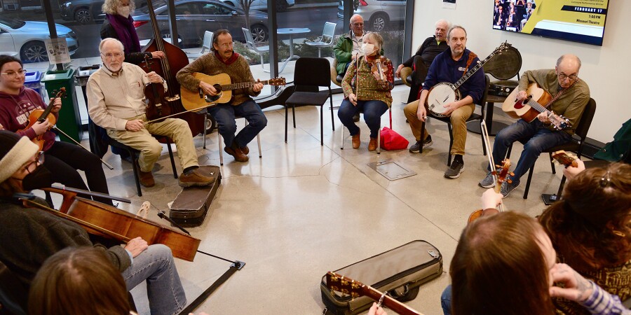 10 musicians sit in chairs in a circle with their folk instruments. An additional musician stands, because she plays the upright bass.