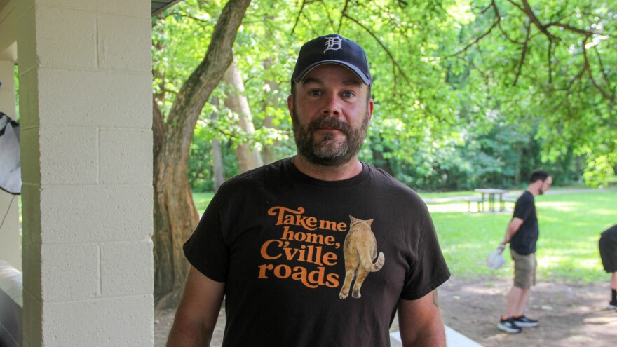 A man wears a shirt that reads "Take me home, C'ville roads," with a photo of an orange cat walking away. 