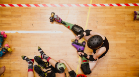 Two roller derby skaters collide while sitting on a wooden-floor rink, wearing helmets, knee pads, and roller skates.