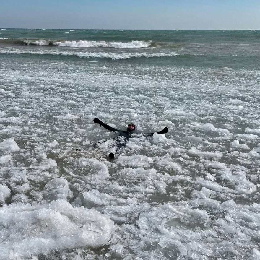 A man in a wet suit floats in a large body of water surrounded by ice.