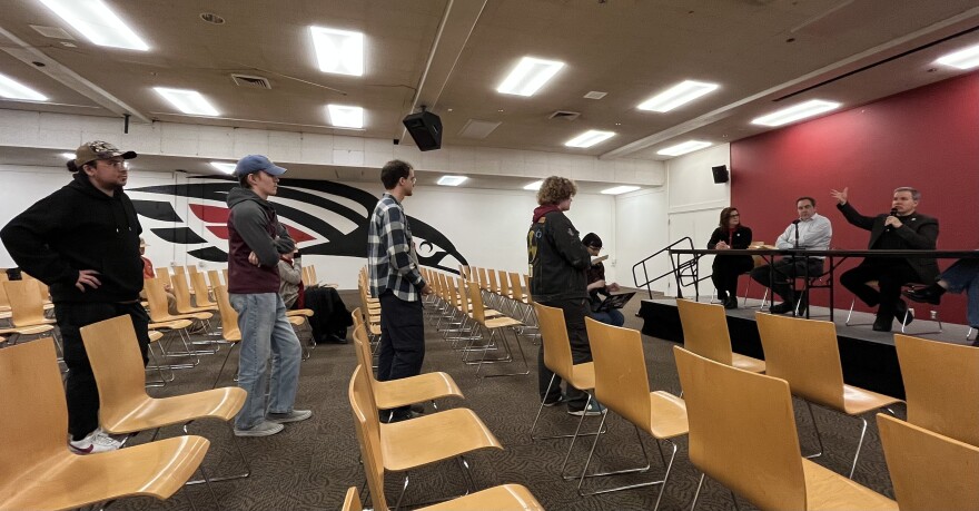Amid rows of wooden chairs, four people are standing in line in a big room. To the right, a man on stage is speaking into a microphone. 
