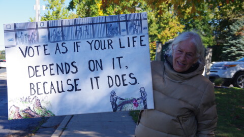 A woman in a tan puffer jacket on Drake Road stands in the shade of a tree holding a white sign that reads in black letters "Vote as if your life depends upon it because it does." 