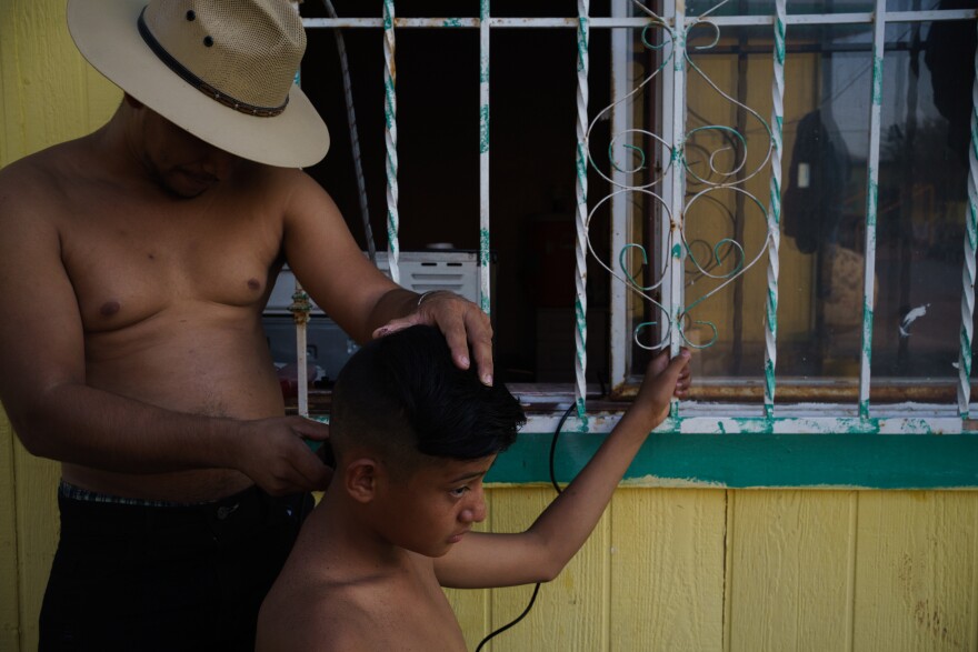 Jose cutting a young man's hair outside the shelter house with clippers plugged-in inside a window.