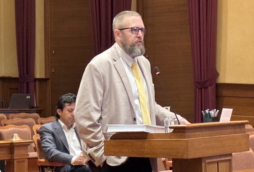 A bearded man in a blazer and tie speaks at a podium in a courtroom, with another man sitting behind him.