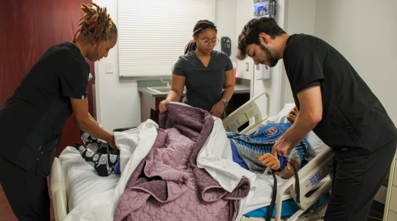 Students attend to the mannequin patient. One lifts the covers near his feet, checking the socks. Another is removing the cover near his stomach. Another is checking a restraint on his arm.