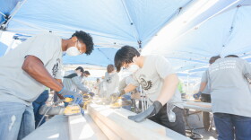 Volunteers wearing masks and safety glasses use power sanders on lumber beneath outdoor canopies.