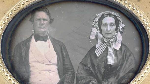 An antique, oval-framed daguerreotype featuring a somber middle-aged man and woman from the mid-19th century. The man on the left has wavy dark hair and wears a dark jacket over a light-colored, double-breasted waistcoat and a stiff white collar with a dark cravat. The woman on the right wears a dark shawl and an elaborate, ruffled bonnet adorned with checkered ribbons that tie in a large bow under her chin. 