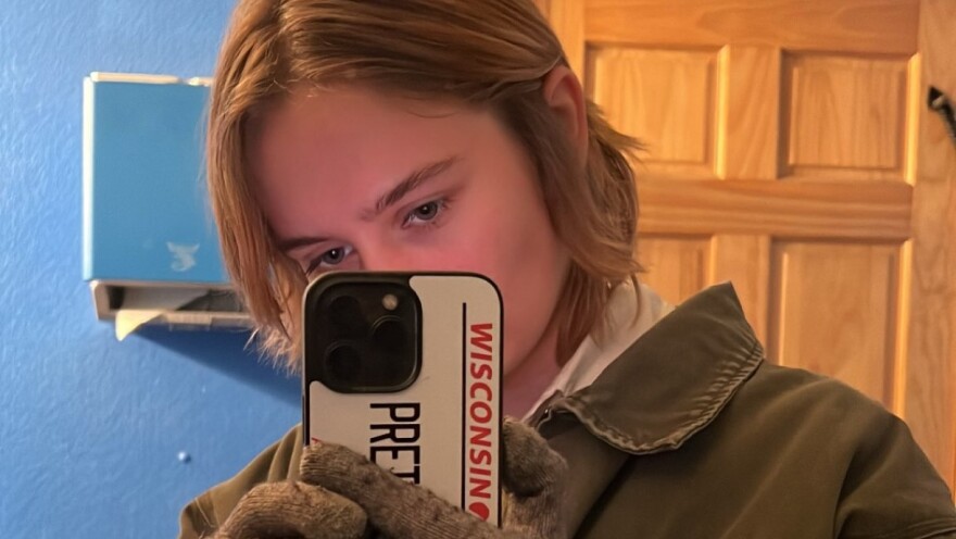 A woman wearing a coat and winter gloves holds a cellphone while taking her photo in a mirror.