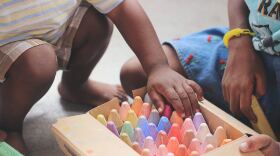 Two Children crouch on the ground near a box of colorful chalk.