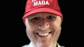 A smiling man is wearing a red hat with white lettering and a white button-down shirt.